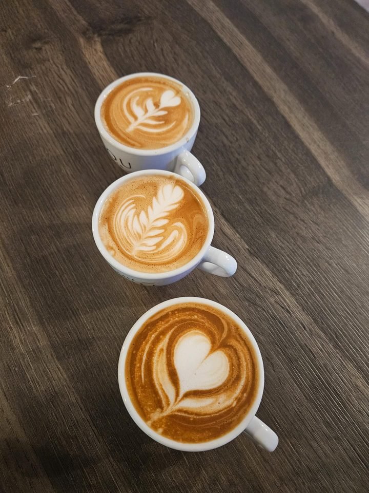 Tres cappuccinos con arte latte alineados sobre la mesa oscura del local.