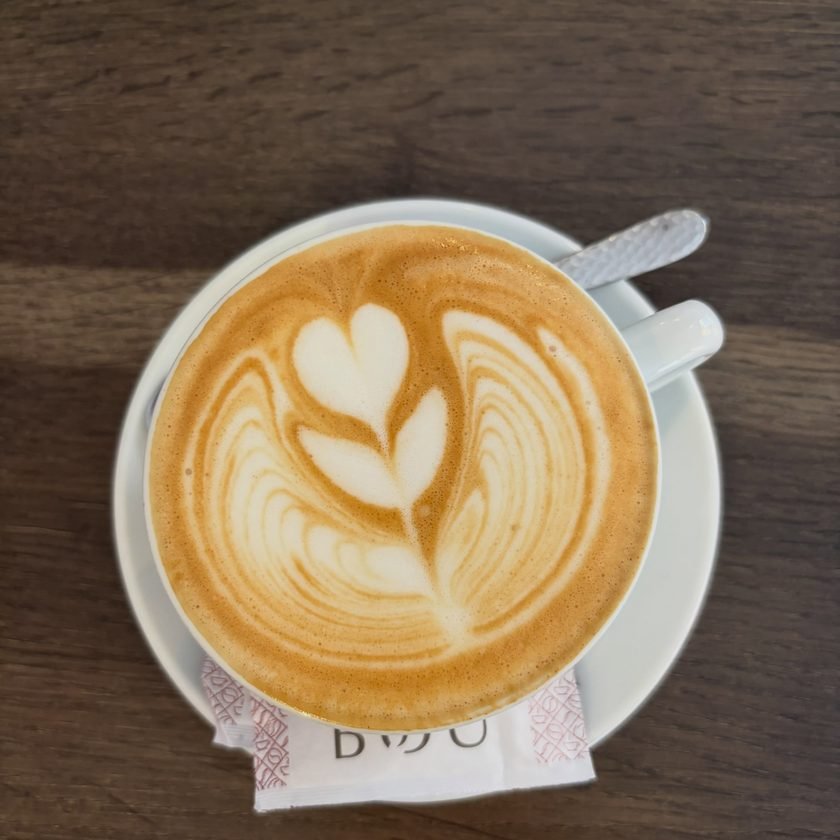 Café con leche con arte latte en forma de hoja, servido en taza blanca sobre la mesa oscura del local.