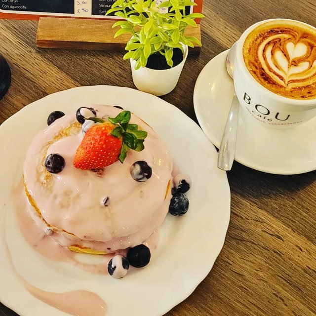 Tortitas con fresa y arándanos junto a un café con leche sobre mesa de madera.
